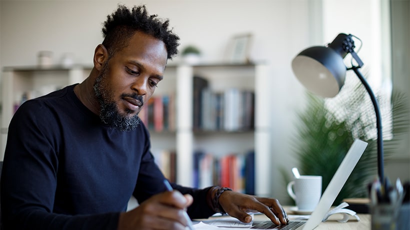Man doing some research on his laptop at a desk and writing notes