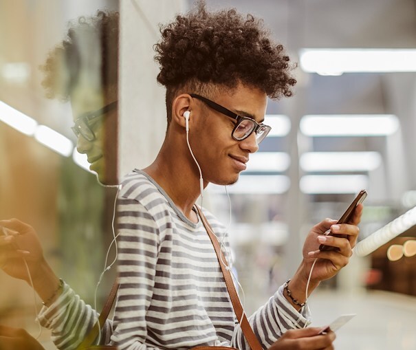 A smiling, young man leaning against a wall and looking at his smart phone